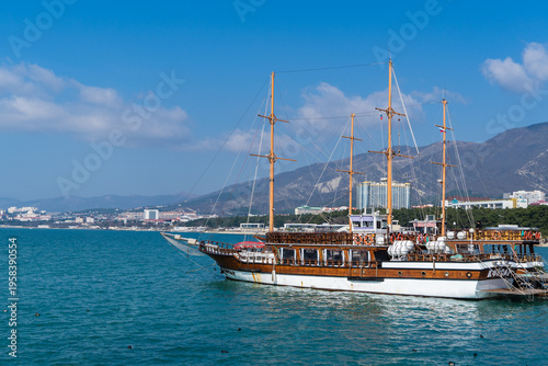 Large, classic wooden sailing ship with multiple masts cruises on calm blue waters, with coastal city and mountains forming scenic backdrop under clear sky. Gelendzhik, Russia - March 2, 2026