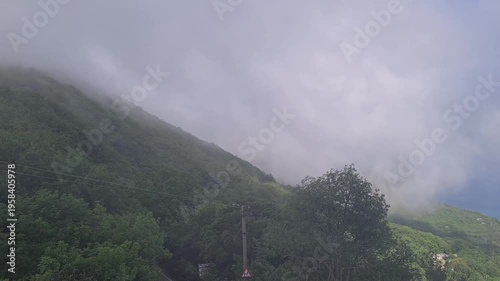 A misty mountain landscape with dense green trees and low-hanging clouds. The scene captures a serene and tranquil atmosphere in nature.