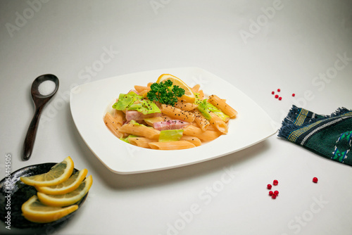 Horizontal minimalist food composition featuring penne pasta with lemon slices, ham, and fresh cabbage, garnished with herbs and served on a white plate. Styled with kitchen props including lemon 