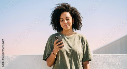Young African woman with curly hair smiles while using a smartphone. She wears a casual green t-shirt against a clear sky background.