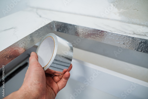 Hand Holding Aluminum Foil Tape Over Appliance Seam, Sealing Duct Edge With Reflective Insulation, Closeup Of Palm And Roll, Textured Metal, DIY Repair Atmosphere, Precise Application