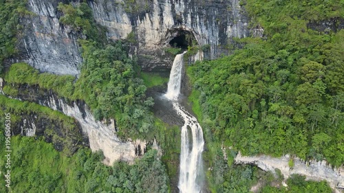 Drone footage of Tisquizoque waterfall pouring from a cave in sheer limestone cliffs, surrounded by dense green forest and a dramatic two-tier cascade.