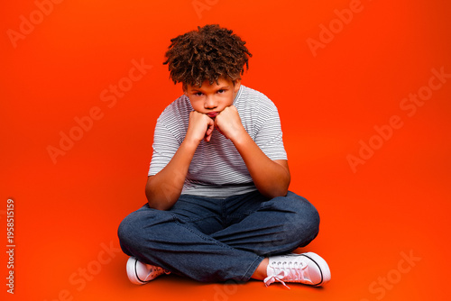Photos Pensive boy sitting cross-legged on an orange background looking bored and upset