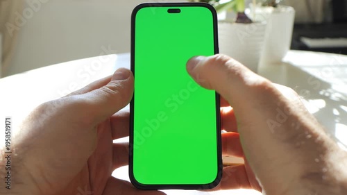 Hands hold smartphone with green screen. Phone held steady, then finger taps screen. Background shows table and potted plant. Natural light illuminates scene
