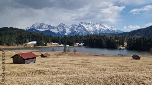 wide-angle landscape of the scenic Geroldsee lake in the Bavarian Alps near Garmisch-Partenkirchen. In the foreground, traditional wooden hay huts (Stadel) sit on a rolling golden meadow.