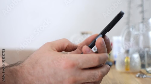 Hand holds black smartphone. Fingers tap screen. Blurred background with glassware. Indoor setting. Neutral lighting. No animals or plants visible