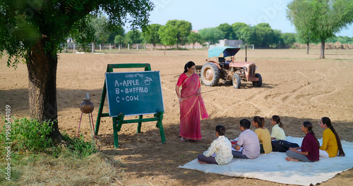 Indian mature adult gen z female lady talk speak teach to villager child day under tree rural area. Happy girl boy group sitting on mat do study look board learn read english at open class field place