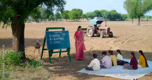 Indian mature adult gen z female lady talk speak teach to villager child day under tree rural area. Happy girl boy group sitting on mat do study look board learn read english at open class field place