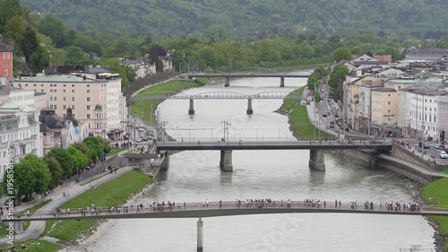 Aerial View of Salzach River in Salzburg Austria with Crowds Crossing Bridges; Scenic Cityscape of Salzburg Old Town featuring Makartsteg Bridge and Alpine Mountains in Summer Establishing Shot