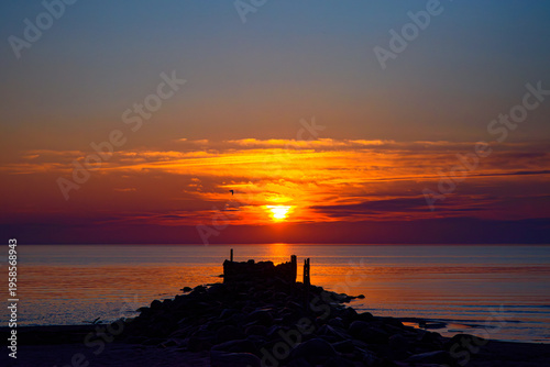 Vivid sunset over calm sea with glowing sky and rocky pier silhouette leading toward the horizon.
