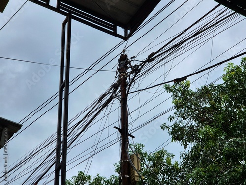 Urban utility pole with tangled electrical wires against cloudy sky and nearby green trees