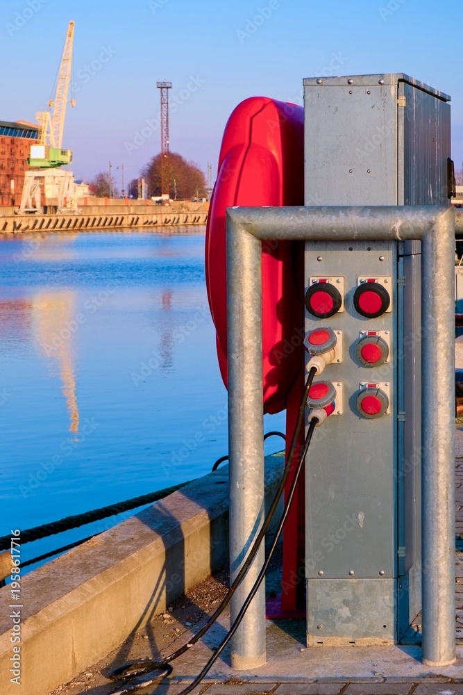 Fototapeta premium Electric shore power pedestal and red lifebuoy at the harbor pier. Port canal with historic brick warehouses and crane. Maritime infrastructure, safety and logistics.