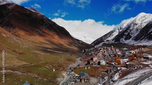drone view of winding river cutting through valley toward scattered houses and towers, golden slopes meeting snowline, atmospheric light and expansive sky create sense of journey and remote mountain