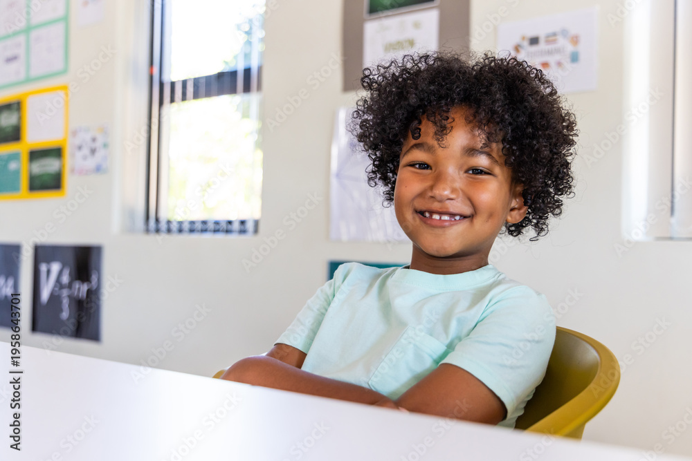 Fototapeta premium African American school-aged boy smiling while sitting in yellow chair at white desk in class