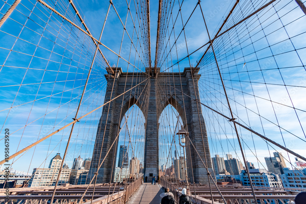 Fototapeta premium Symmetrical view of the Brooklyn Bridge pedestrian walkway leading toward the stone tower, framed by suspension cables and city buildings under a clear blue sky in New York City, USA. 