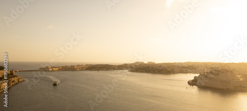 The Grand Harbour glows under a warm sunset, with Fort St. Angelo standing as a sentinel over the tranquil waters as a ferry crosses the historic port.