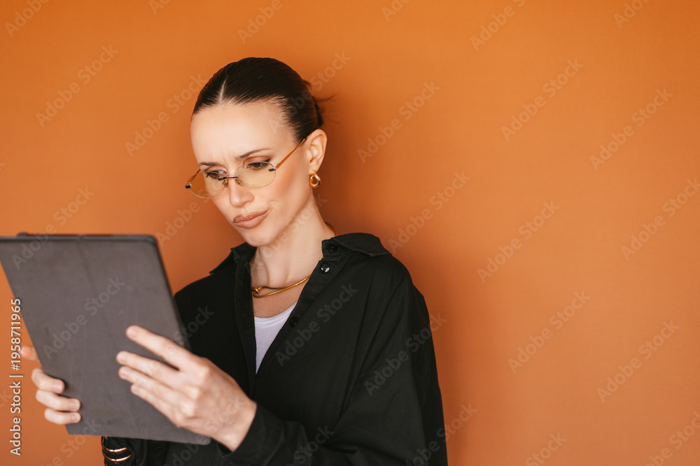 Fototapeta premium Serious young female trader in rimless glasses holding and focused reading a digital tablet. Doubting professional analyzing financial market data, crypto or stock charts. Confident girl.