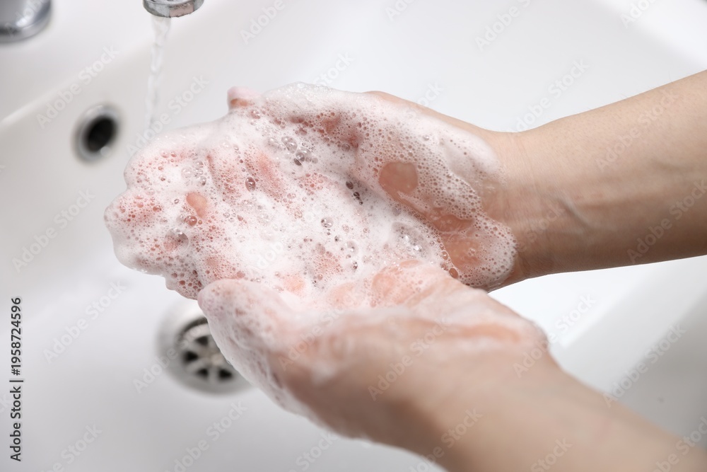 Fototapeta premium Woman washing hands with soap above white sink, closeup