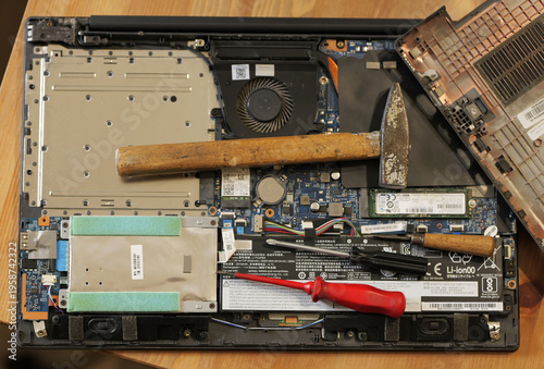 Photos An opened laptop with exposed components lies on a wooden surface, with a hammer