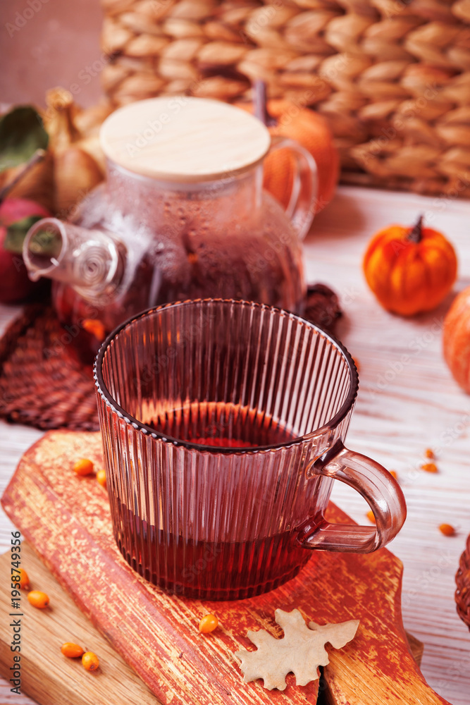Fototapeta premium Glass cup with red herbal tea and teapot placed on rustic wood with autumn foliage, apple and pumpkins in background