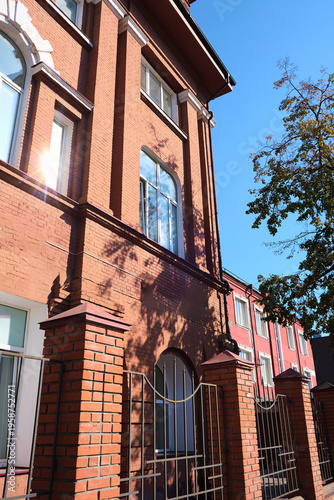 Facade of an old red brick building with arched windows and decorative white accents, partially shaded by trees and sunlight reflections. The image is suitable for topics about building restoration