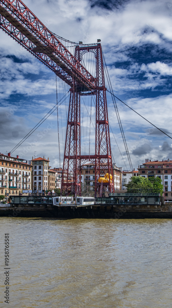 Fototapeta premium Puente de Vizcaya sobre la ría del Nervión, icónico puente transbordador de estructura metálica roja que une Portugalete y Getxo, bajo un cielo azul con nubes dinámicas en Bizkaia.