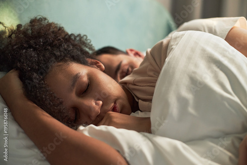 Young adult Black woman and young adult Caucasian man sleeping closely together in bed, both resting peacefully with heads on pillows, intimate moment showing couple embracing