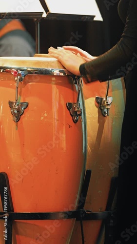 Close up of a percussionist's hands on a vibrant orange conga drum. Warm stage lighting highlights the instrument and the artist during an energetic live music performance at a jazz festival
