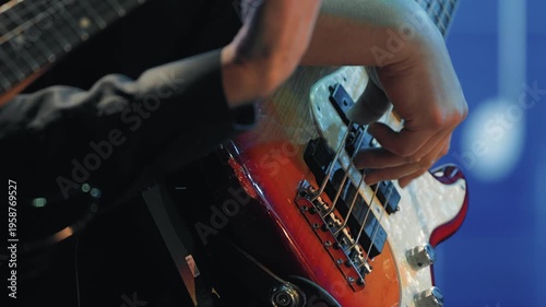 A tight shot of a musician's hands playing a sunburst bass guitar. The scene is lit with warm stage lights against a blue background, capturing the energy of a live jazz performance