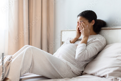 Tableau sur toile Pregnant Black woman lying in bed covering her face with hands, expressing stress or headache