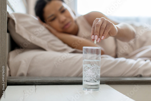 Young african american woman lying in bed taking medicine with glass of water, feeling unwell, representing illness, treatment and healthcare routine at home environment