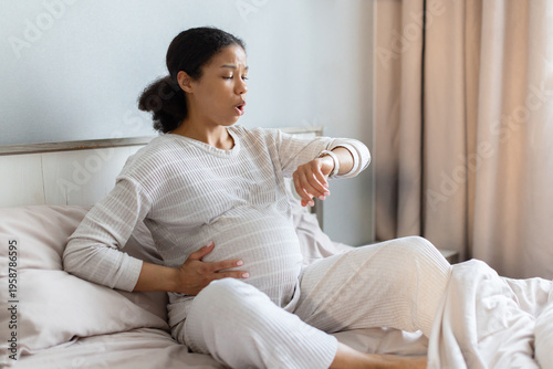 Pregnant Black woman sitting on bed timing contractions, checking watch with focused expression. Labor preparation concept in bright cozy bedroom