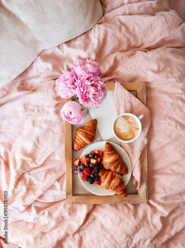 Overhead flat lay of a Mother's Day breakfast in bed featuring croissants, fresh berries, coffee, and pink peonies on a soft pink bedspread