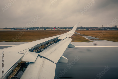 Airplane Wing With Flaps Extended During Rainy Takeoff Over Wet Runway At Cloudy Airport
