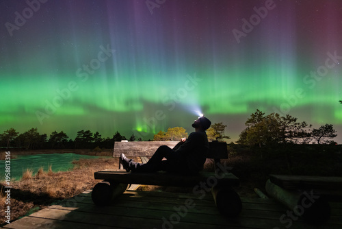 A man sitting on a wooden bench watching the spectacular Aurora Borealis green and purple lights in Estonian bog.