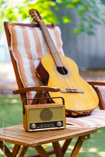 Cozy outdoor relaxation space with wooden garden chair, acoustic guitar and vintage radio placed on small table. Peaceful backyard leisure scene for music, rest and summer recreation in green garden.