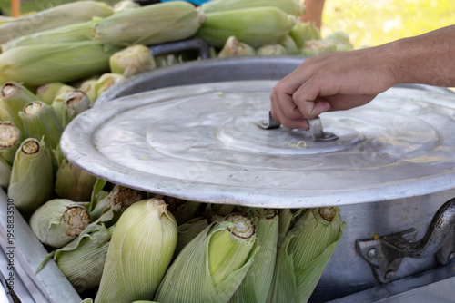 A hand lifts a pot lid, revealing steaming corn on the cob. 