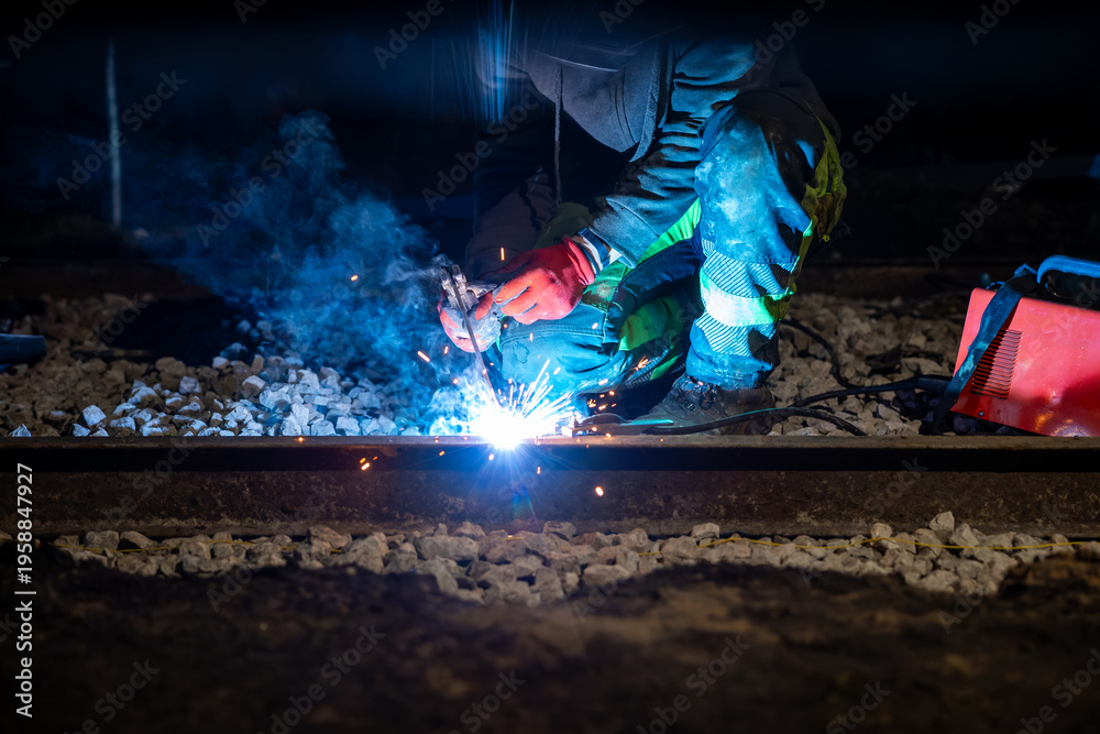 Fototapeta premium A worker in reflective gear arc welds a steel rail at night on ballast, sparks and blue smoke rise, portable welder nearby, task lighting isolates gloves and stripes.