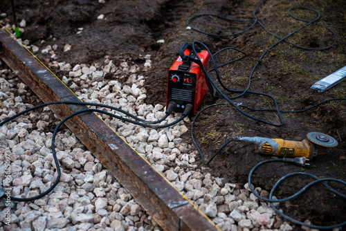 Fotografi A red inverter welder sits on compacted dirt by crushed stone ballast, leads on a rusted rail along a chalk line, with an angle grinder nearby under overcast light