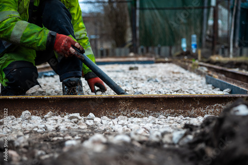 Foto A worker in a green jacket and red gloves kneels by exposed rails, prying steel with a crowbar as crushed stone ballast extends