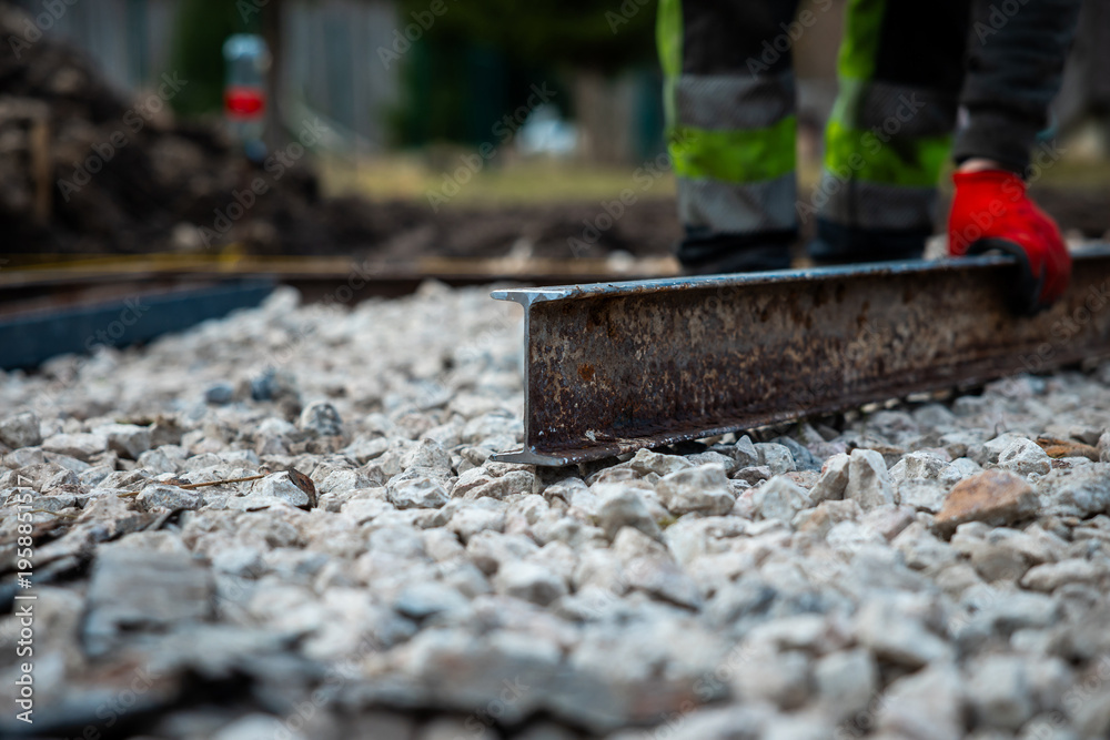 Naklejka premium A worker in red gloves and high visibility pants drags a steel straightedge over crushed stone, daytime setting, shallow depth of field emphasizes precision and grit.