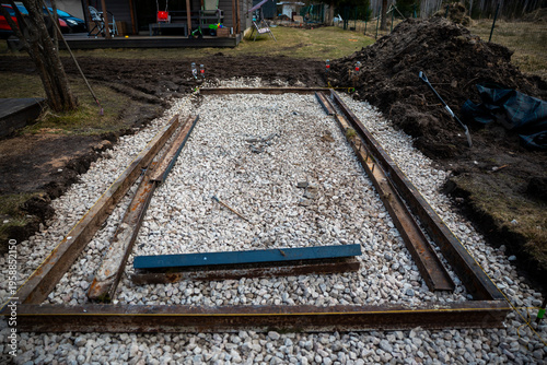Rectangular trench filled with crushed stone sits beside a house deck. Rusty steel rails and string lines mark layout, with shovels and levels present in cool daylight.