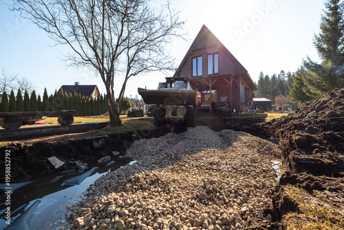 A skid steer dumps crushed stone into a trench by a dark wood A frame cabin with tall triangular windows and a wraparound porch, under springlike light with wet soil and water.