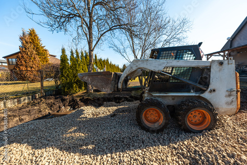 Bild auf Leinwand A compact skid steer with orange wheels raises a bucket on a gravel bed near a residential fence, arborvitae, and leafless trees, grading and excavating in cool daylight
