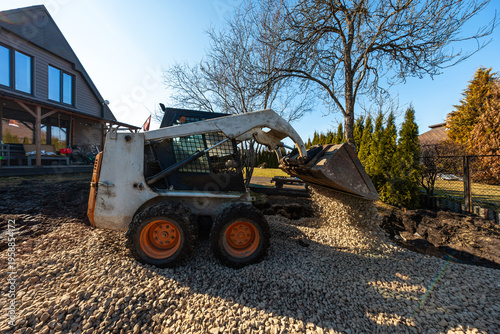 A compact skid steer pours light crushed stone onto a yard beside a modern wood sided house, with chain link fence, tall evergreens, bare branches, and crisp afternoon shadows.