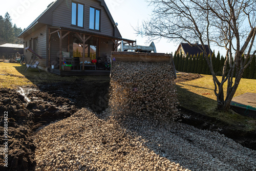 A front loader tips crushed stone into a dug path by a timber clad house with porch. Pebbles fly mid air on a sunny day, curving route past a bare tree and hedges.