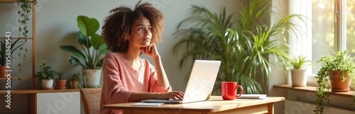 Curly haired woman types on laptop at wooden desk with red mug and green plants. She looks thoughtfully out window. Natural light fills cozy room. Home office setup for remote work.