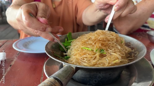 Elderly East Asian woman mixing Casseroled Shrimps with Glass Noodles.