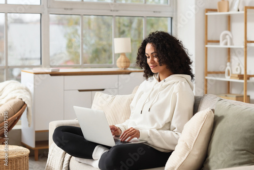 Foto Young African-American woman working with laptop on sofa at home
