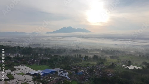 Aerial sunrise view of a misty valley with scattered villages, lush greenery, and a distant mountain range under a glowing sky